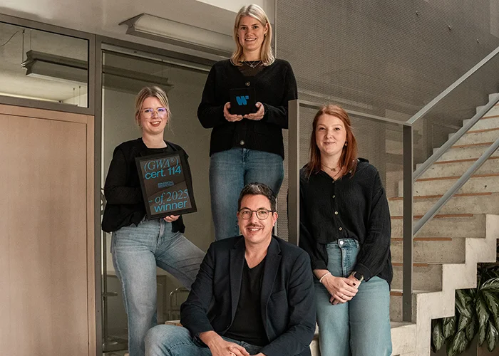 Vier Personen posieren im Innenbereich eines Büros auf einer Treppe, zwei halten Auszeichnungen in den Händen, alle blicken in die Kamera.