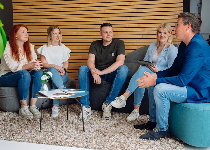 Fünf Personen sitzen auf Sofas in einem Besprechungsbereich, sie unterhalten sich miteinander, ein Mann spricht und gestikuliert, die anderen hören zu.