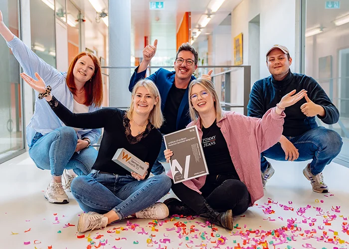 Fünf Personen sitzen und knien in einem Büroflur, halten Auszeichnungen und strecken die Arme hoch, während buntes Konfetti auf dem Boden liegt.