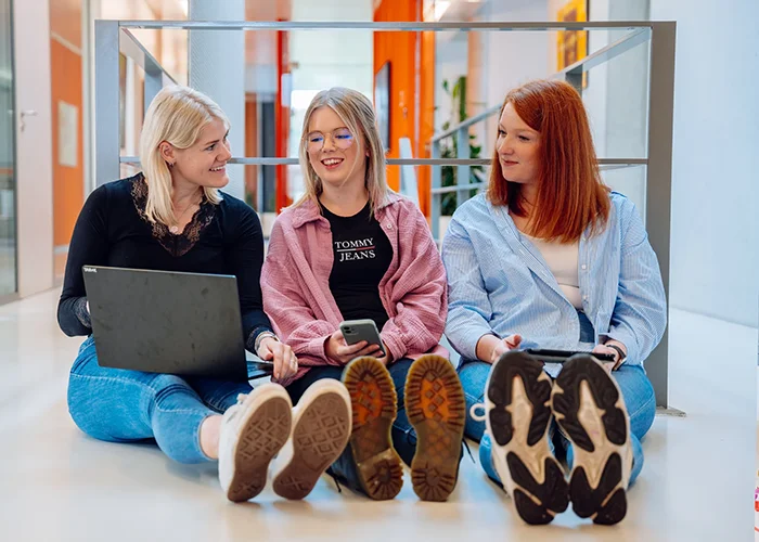 Drei Frauen sitzen auf dem Boden in einem modernen Büroflur, eine arbeitet am Laptop, eine hält ein Smartphone, alle schauen sich lächelnd an.
