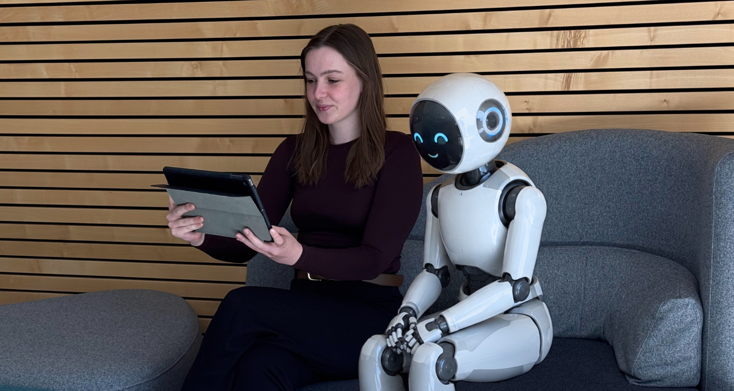 Frau sitzt mit freundlichem Roboter auf einem Sofa und schaut gemeinsam auf ein Tablet